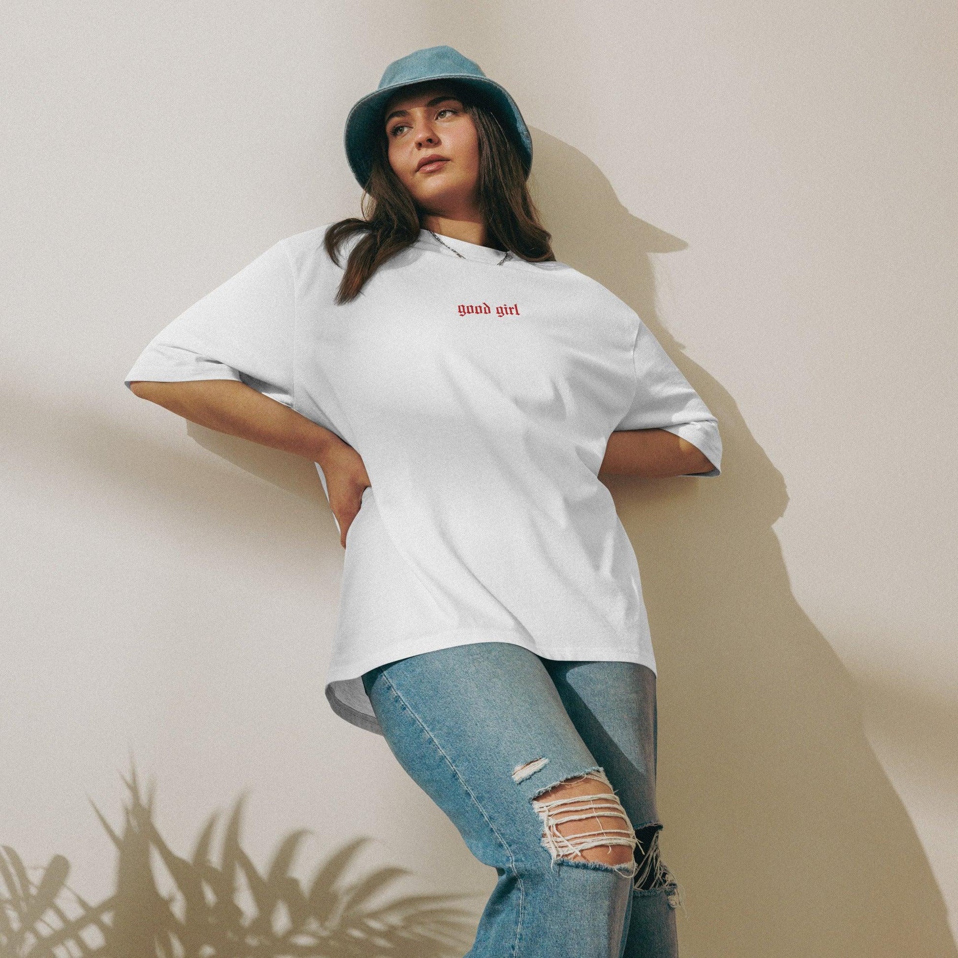 Woman wearing embroidered good girl tee in white with red text paired with ripped jeans and a blue bucket hat